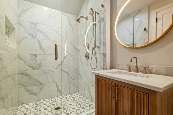 Marble-look tile shower beside a fluted wood vanity and a round lit mirror