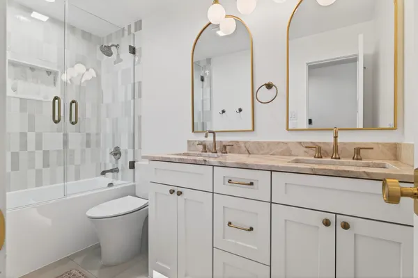 Bath with a white vanity, a stone top, brass arched mirrors and a glass tub enclosure in white tile
