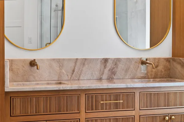 Stone vanity top and backsplash with wall-mount brass faucets under oval mirrors
