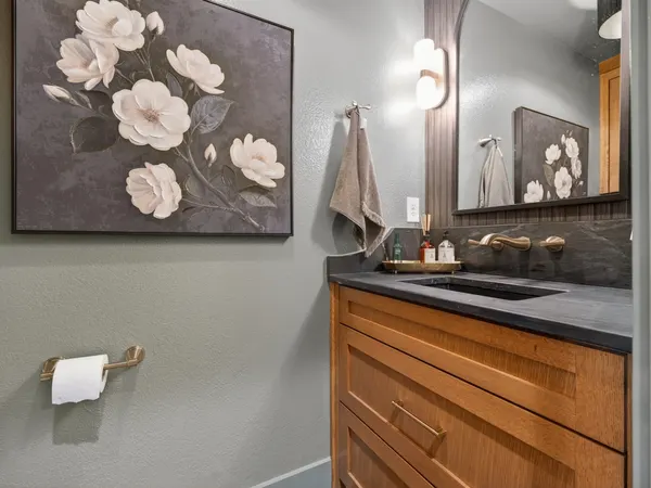 Powder room with a wood vanity, a dark stone top and backsplash, and a brass wall-mount faucet