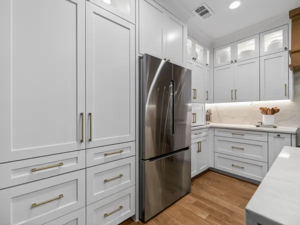 White cabinetry with brass pulls around a stainless refrigerator, with lit glass-front uppers