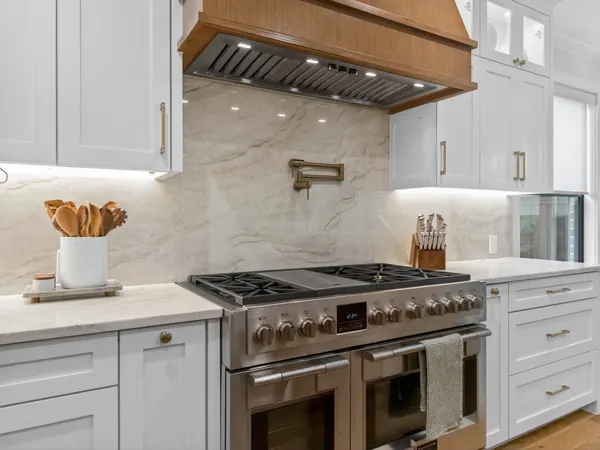Professional-style range and brass pot filler under a wood range hood, against a slab backsplash