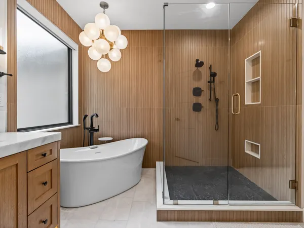 Freestanding tub and glass shower against fluted wood-look tile, under a glass globe chandelier