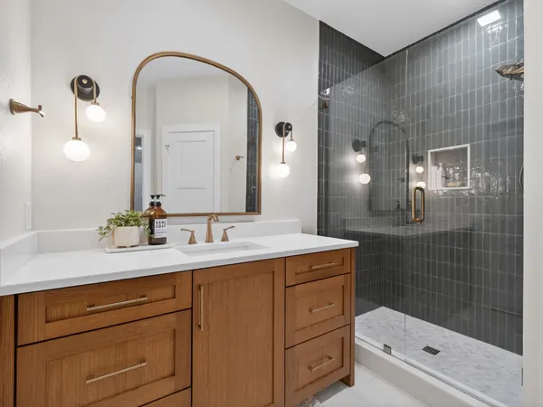 Bath with a wood vanity, a brass arched mirror and a glass shower in dark green tile
