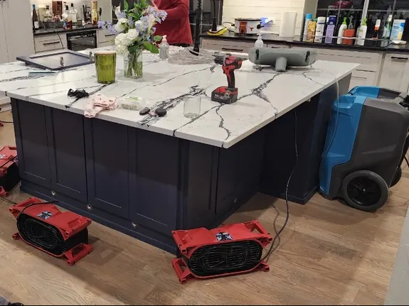 Air movers and a dehumidifier drying the wood floor around a navy kitchen island with white marble-look counters